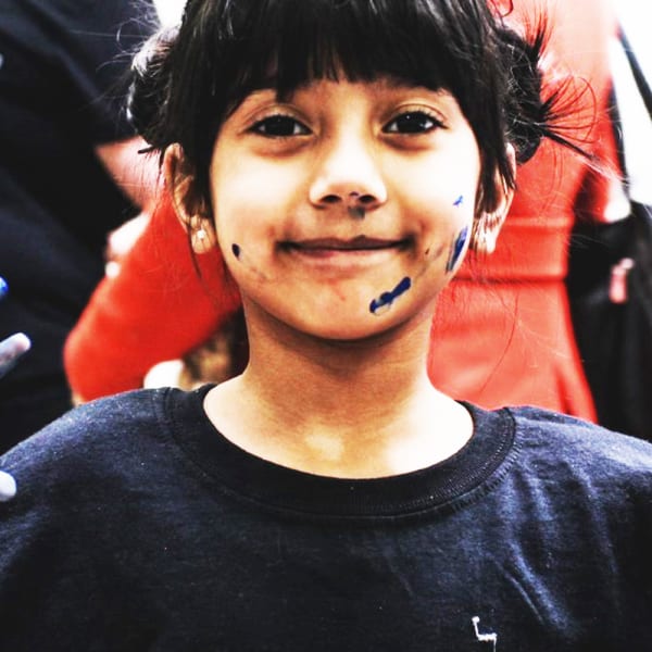 A Young girl holds up her hands showing that they are covered in blue paint, she also has a few paint marks on her face.