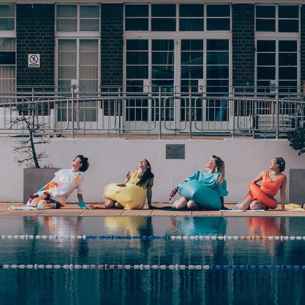 5 adults pose in brightly coloured outfits by an outdoor swimming pool