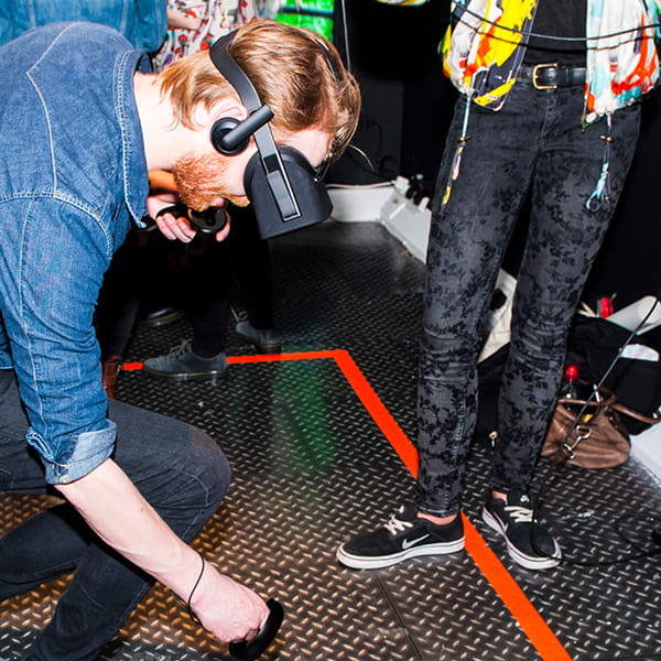 Image of man in denim shirt and jeans wearing a virtual reality headset while looking and bending down within a red rectangle taped to the floor.