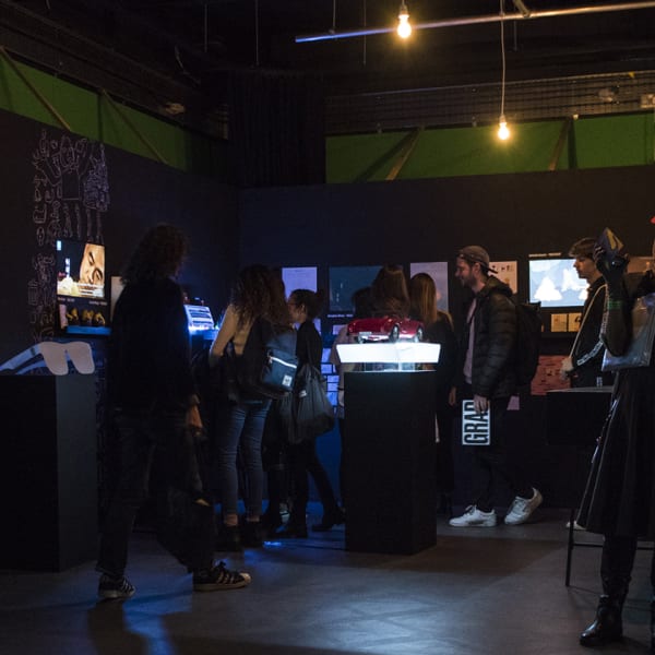 Exhibition shot of a dark space with visitors watching screens and interacting.