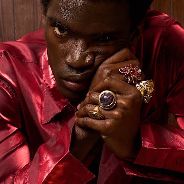 A man looks at the camera. He wears a silk pink shirt and raises his hands up into clenched fists. 