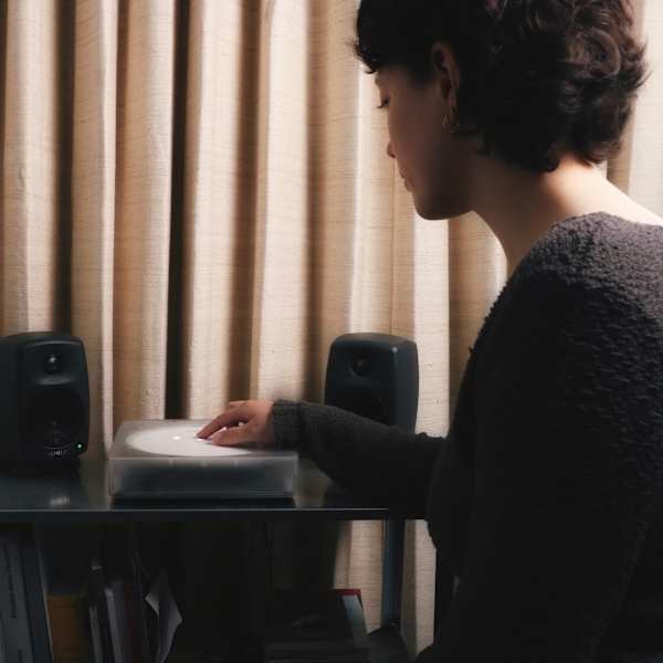 A person is interacting with a small digital turntable on a stand, with speakers on either side. There is a soft draped curtain behind and a lamp stand just visible to the left.