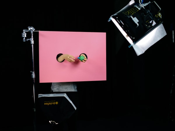 Two hands emerging through hole in a pink screen holding a green item