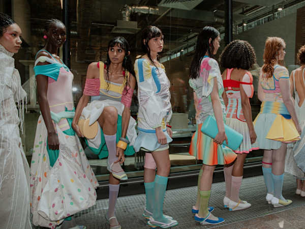 Shot from the side, a long row of models in colourful sheer and knitted garments referencing retro sportswear are lined up ready to go on the runway.