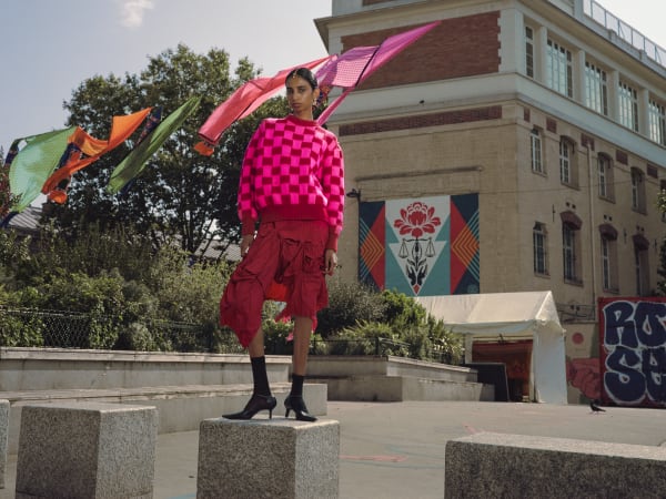 A model in bright pink skirt and patterned jumper is standing on a square marble platform next to a large commercial building just visible. There is greenery in the background and colourful gingham fabrics hanging on a line overhead.