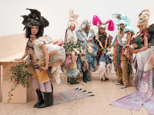 Models in avantgarde garments in muted and pastel colours are posing backstage. All are wearing headdresses and there are woven and plaited textiles and elements, patterns and tree branches incorporated.