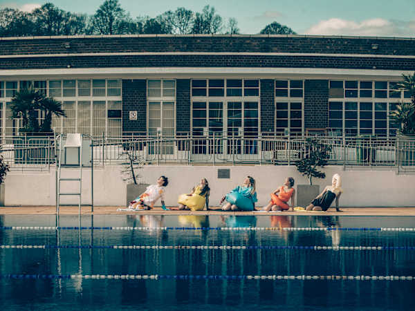 5 adults pose in brightly coloured outfits by an outdoor swimming pool