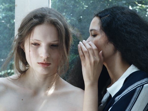 A fashion portrait with two models, one facing the camera, the other has her head turned towards her and is whispering behind her hand. We can see greenery via a pane in the background.