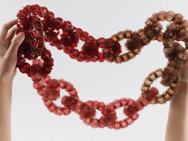 Hands holding up a red, beige, dark and amber-coloured interlinked sculptural necklace made of delicate natural materials.