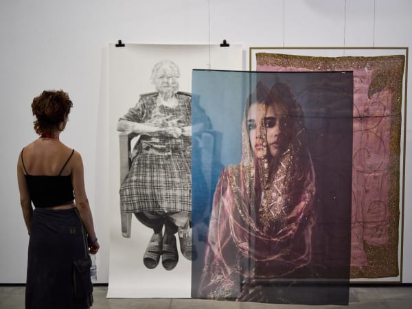 An observer is standing looking at a trio of large-scale hanging photographic, drawing and textile artworks, the photo is a stylised shot of a woman in a veil, next to a pink embroidered sari cloth with gold trims. Behind this is a detailed drawing of a seated woman with crossed legs wearing tartan. 
