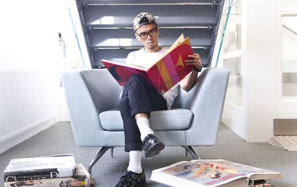Photo of man sitting in a chair under the stairs reading a large pink, yellow and red book