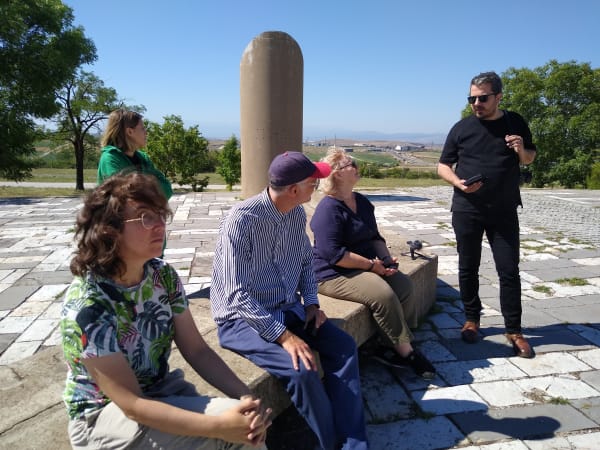 Five people in a paved outdoor space.  Three are sitting on a concrete bench and talking with a person standing.  Behind them is a small concrete monolith and in the background is a rural landscape.