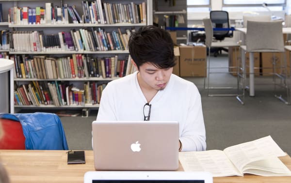 Photo of a man siting at a table in the library using his laptop and reading a book