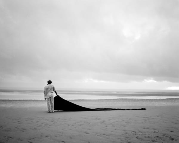 Black and white photo of a figure standing on the beach holding a swathe of black fabric.
