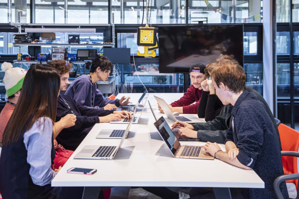 A group of 6 students sitting around a table with laptops on it. They are talking to each other and looking at their devices. In the background is a set of shelves with various vintage computers, radios and other technology.