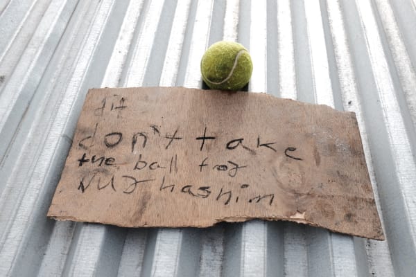 Wooden board with a handwritten sign and a tennis ball on a metal fence