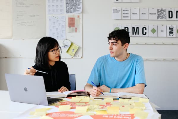 Two people are sitting together at a table. On the table is a poster covered with post-it notes and a laptop. They are discussing the project in front of them.