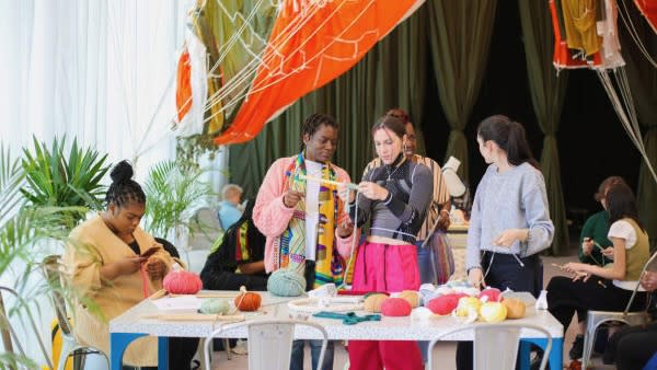 People in a room, one sitting down knitting, two standing up chatting, surrounded by brightly coloured knitting yarn.