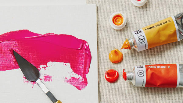 Two tubes of open paint, with a canvas with pink paint smeared on, next to a paintbrush. 