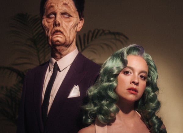 Tall male model with prosthetic face standing beside female attractive female model with retro hair and dress.