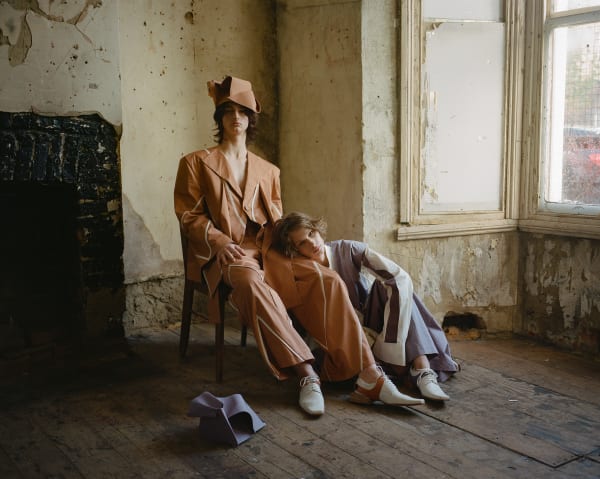 Male models in tailored outfits sitting in dilapidated room.