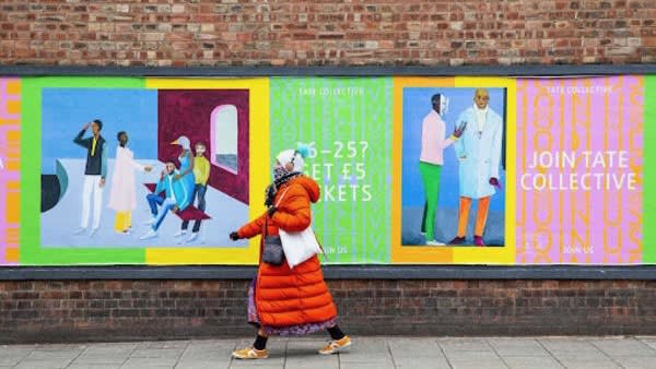 Person walking along a street next to a wall with colourful Tate advertising posters lined across it.