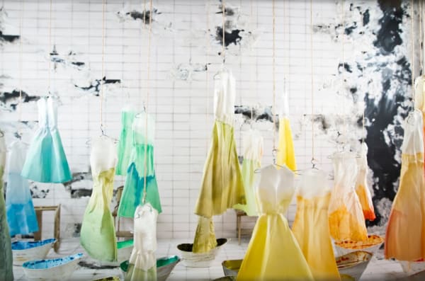 multiple hanging dip-dyed dresses in front of a cermaic tiled and cloud decal backdrop. Dresses all white at the top, and dyed in various colours at the botom- blue, teal, green khaki, yellow and orange.