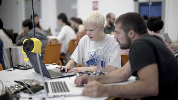 2 students working on laptops in the foreground. In the background there are other students working at other tables. 