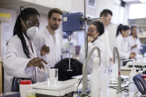 Cosmetic Science students from London College of Fashion in a lab.In the foreground a person is pouring clear liquid into a glass beaker. 