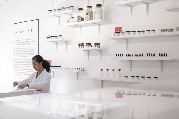 Woman in white lab coat with shelves of cosmetics behind her. LCF Formulates Cosmetic Science exhibition, Fashion Business School Summit, Kingly Court. Copyright holder: Emmi Hyyppa