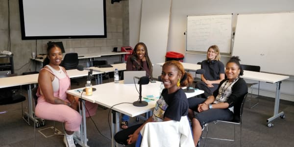 5 people sat round a table with microphones and headphones. They are all looking at the camera and smiling. 