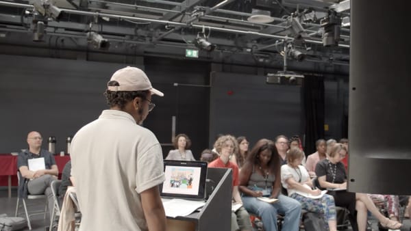 A man stands in front of a podium in front of a seated crowd giving a speech at Wimbledon College of Arts