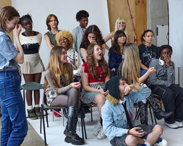 A group of students come together in a group, some sitting some standing. They are in a photo studio, some of them are laughing at something out of shot