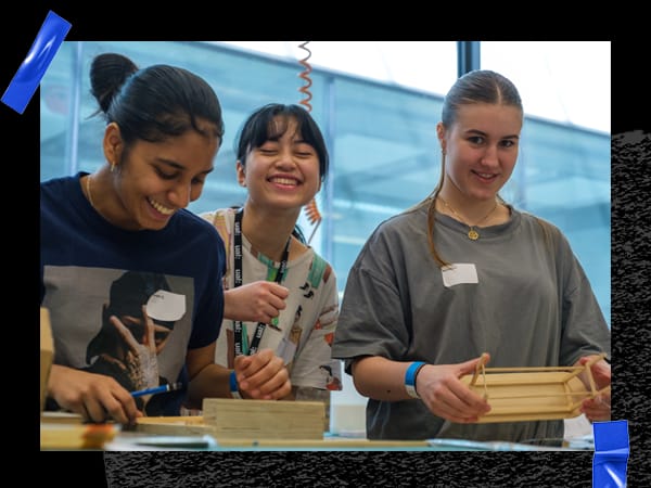 3 students standing together in a workshop, smiling at the camera and holding scale models of furniture