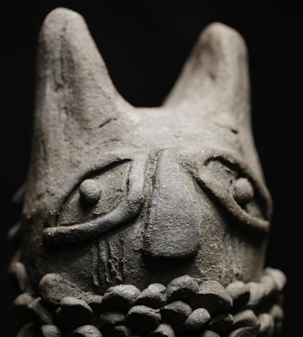 A dark grey clay figure shot against a black background.  It has exaggerated eyes, a prominent nose, and layered, scale-like textures around its base.