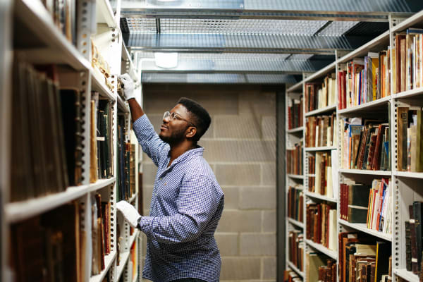  A man in a checkered shirt and white gloves reaches for a book on a tall shelf in a library archive.