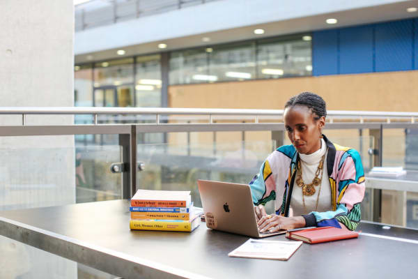 Woman at a desk looking at her laptop with a pile of books next to her. MA Arts and Cultural Enterprise, Rochelle Saunders in the studio. Copyright holder: Alys Tomlinson
