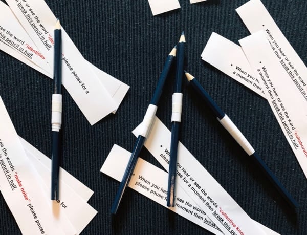 Pencils and slips of paper on a black table. The slips of paper feature instructions which are partly covered by the pencils. These include, “when you see or hear the word collaboration…please pause for a moment…then break the pencil in half.”