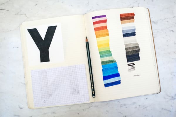 Lined sketchbook open on table with a pencil, displaying large letter Y and test colour stripes.