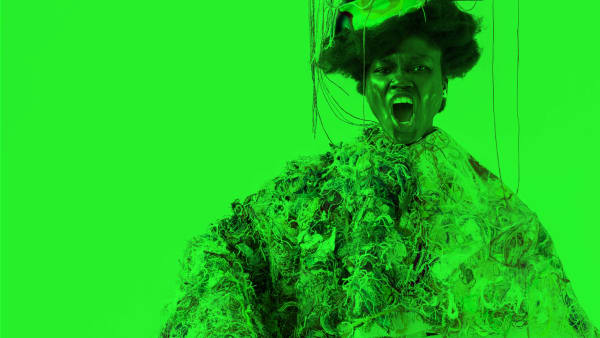 A person shouts at the camera while wearing a tunic covered in multi-coloured strings and material. They also have a monitor on their head, which is covered in multi-coloured string