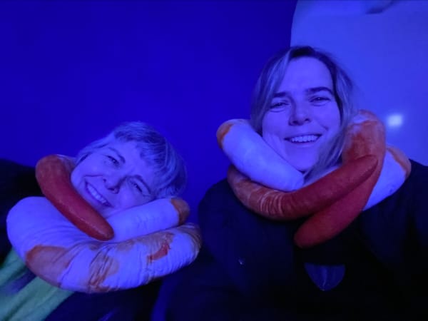 A purple hued photo of two women smiling at the camera, there are snake-like pillows wrapped around their necks