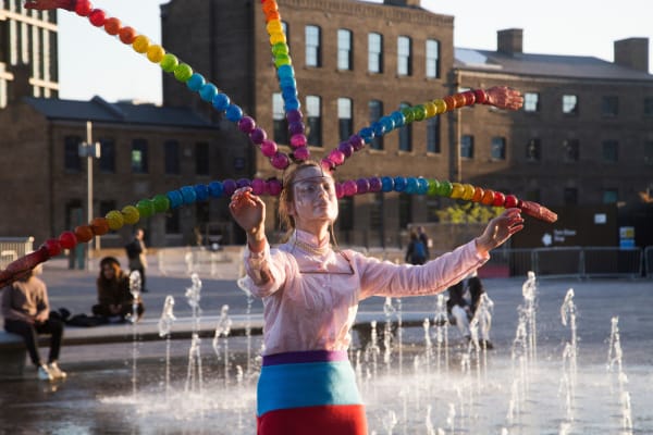 Performer with a large headpiece of colourful balls and a translucent face mask