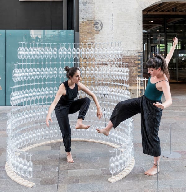 Two people mimicking each other's balance surrounded by a semi circular tower of wine glasses