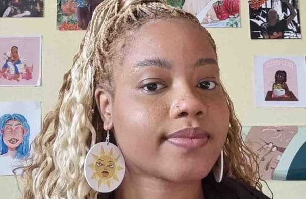 Emmanuelle looks directly into camera, they are wearing earrings with the sun on, have blond braids and a nose ring.
