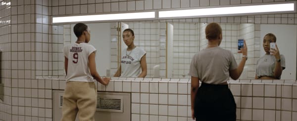 Two people looking into a mirror in a public toilet, one taking a selfie on a phone