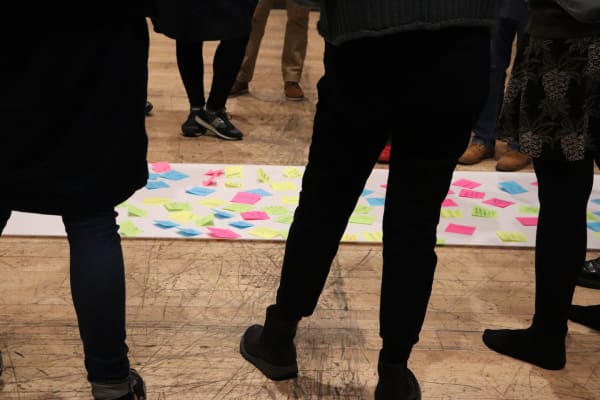 A detail shot of people standing around a piece of paper on the floor with coloured post-it notes attached to it. The photograph only shows the people from the waist down.