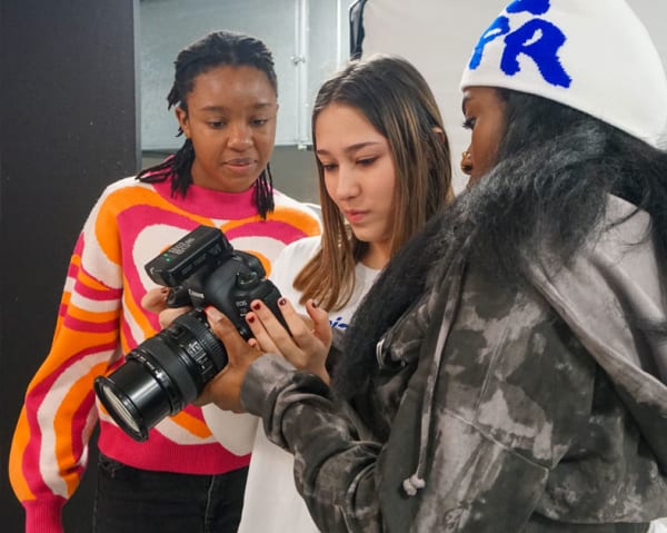 3 students stand together looking at photos taken on a camera. 