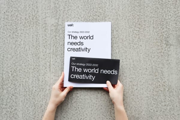 Person's hands holding a printed booklet and leaflet against a concrete wall which is in bright natural light. The leaflets read 'UAL, our strategy 2022-2032, The world needs creativity'.