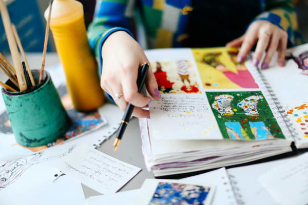Woman's hands with purple nail varnish flipping through brightly painted and collaged sketchbook. Jar of paintbrushes on the left.