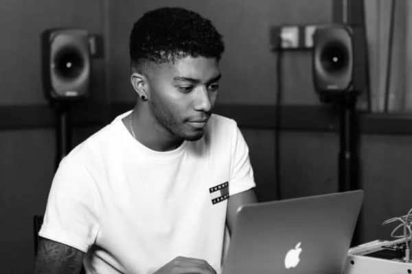 A man sits at an apple laptop looking at the screen. He is wearing a white t-shirt and there are two speakers visable behind him